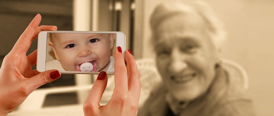 A phone's screen shows a baby with a pacifier, while a photo is being taken of a smiling old woman, symbolizing a person's life journey from infancy to the present.